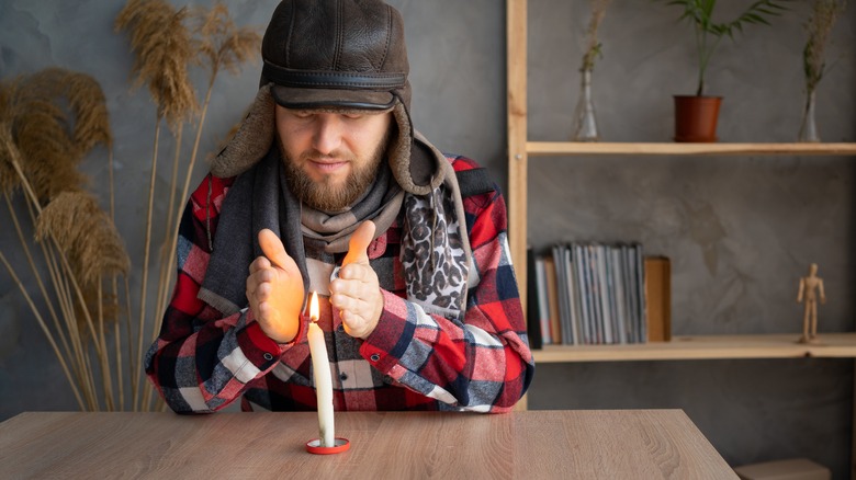 A bearded man dressed in a warm winter hat and a scarf sits at home at a table and warms his hands from a burning candle