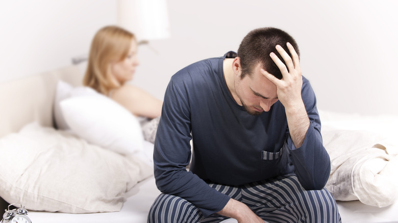 A frustrated man sitting on the edge of bed while his partner sits in bed