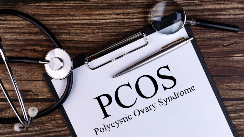 a clipboard that says PCOS polycystic ovary syndrome on a wooden surface with a stethoscope, a magnifying glass, and a pen