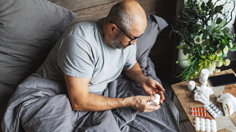 A man in bed looking at medicine bottle with a nightstand of many medications