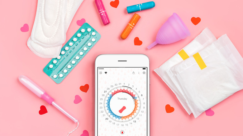 Phone with period tracking app open on a pink table surrounded by menstrual hygiene products