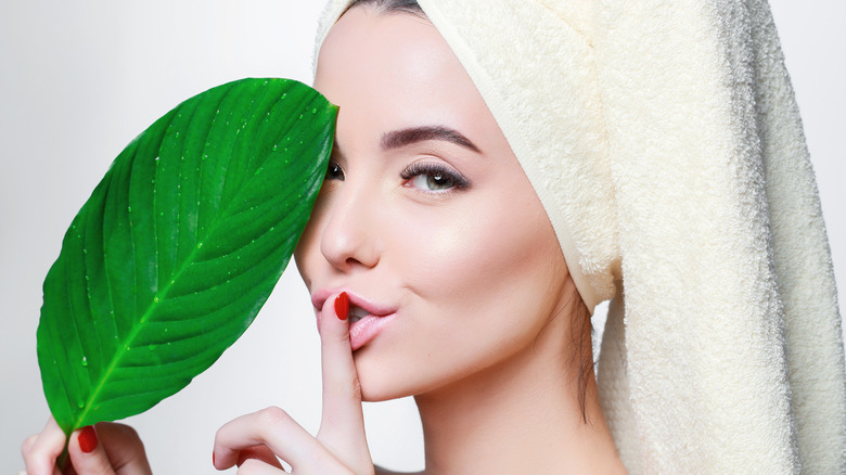 Woman with healthy glowing skin and hair wrapped in a towel holds green tropical leaf and has her index finger held up to her lips as if to say "shhh". . 