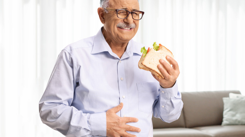 An elderly man experiencing pain while eating a sandwich