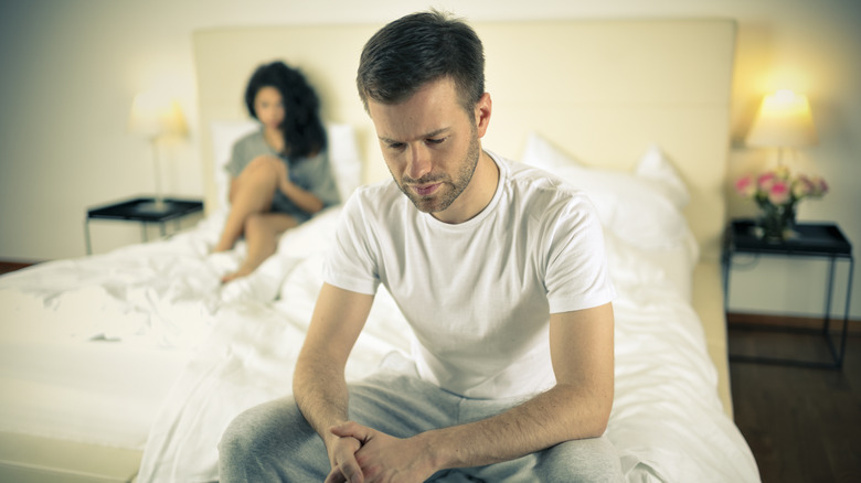 A man sitting on the edge of the bed with his frustrated partner in the background