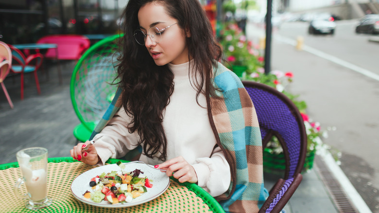 Young woman sitting at cafe table wrapped in a sweater and blanket while eating a salad