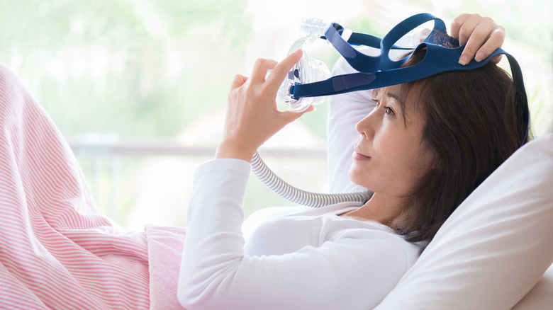 Woman in white clothes lying in bed putting on her CPAP mask 