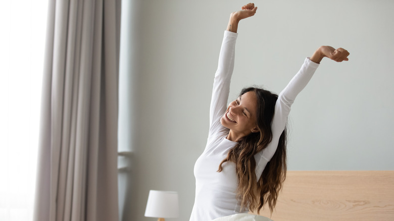 Smiling woman in white nightwear sitting in bed stretching her arms after sleep 