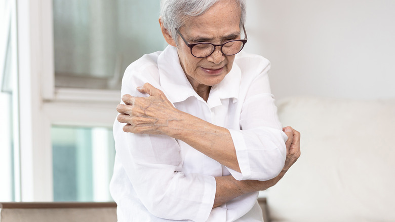 Older woman in glasses sitting on bed scratching her body