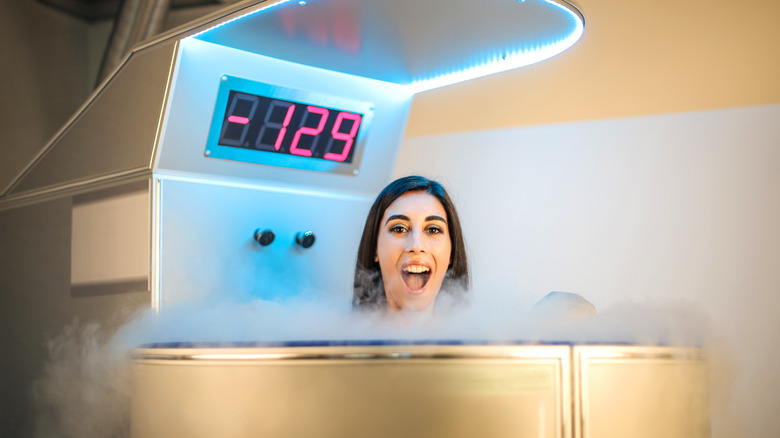 Woman with long brown hair in a metal cryotherapy chamber with her mouth wide open and liquid nitrogen coming out of the tank around her; temperature reading says -129 in red letters