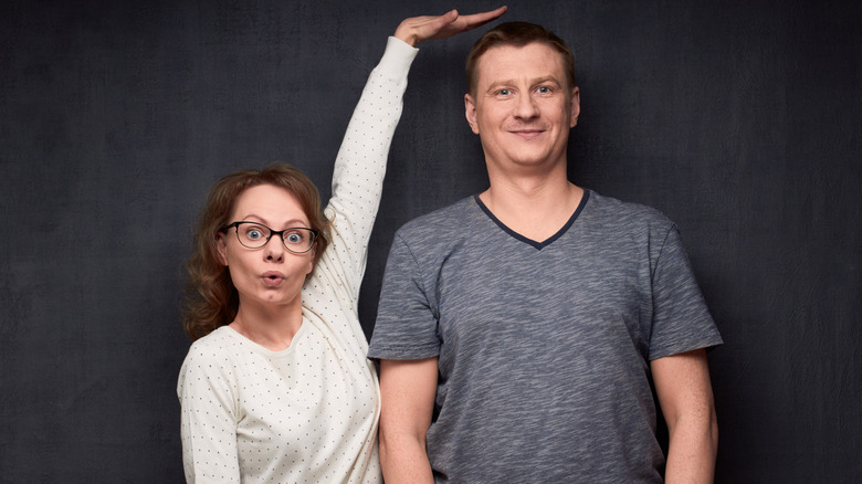 a surprised looking woman measures a much taller man with her raised hand above his head