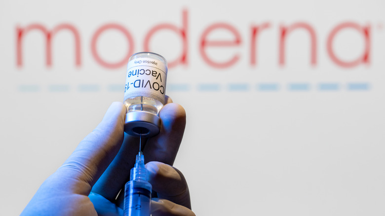 Gloved hand holding vial of COVID-19 vaccine and syringe against "Moderna" sign in background