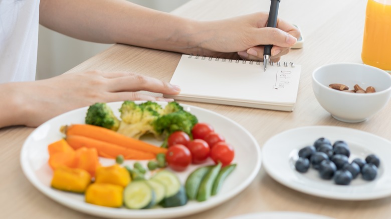 fresh fruits and vegetables on a table with a female writing a dietary plan