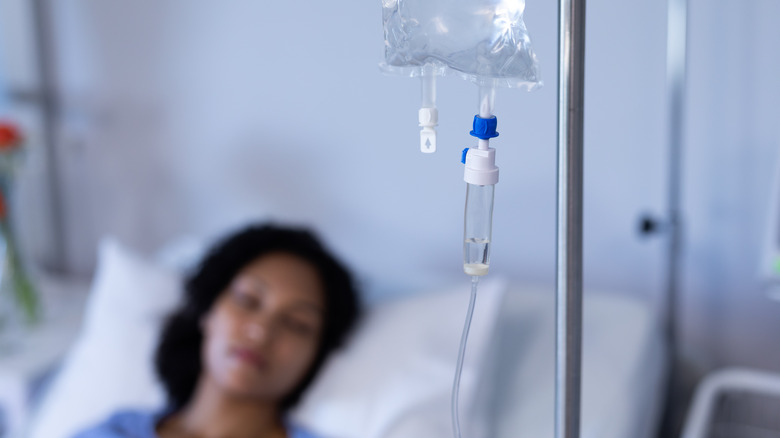 Woman lying in hospital bed in background with IV stand and bag in focus in the foreground