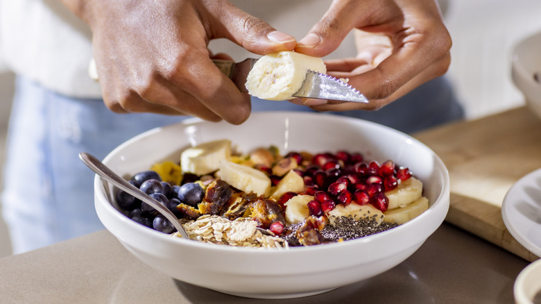 A man's hand slicing a banana over a bowl of oatmeal with fruit