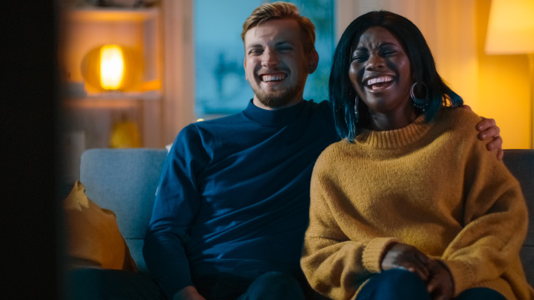 Couple sitting on couch in dark room laughing and watching movie