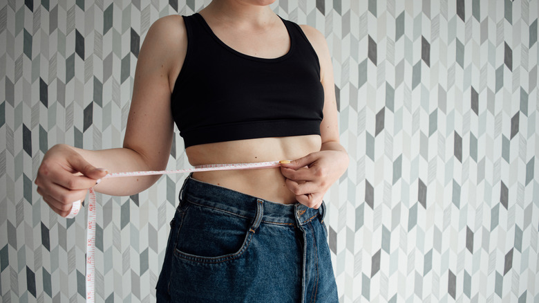 a woman measuring her waist wearing jeans and a black sports bra 