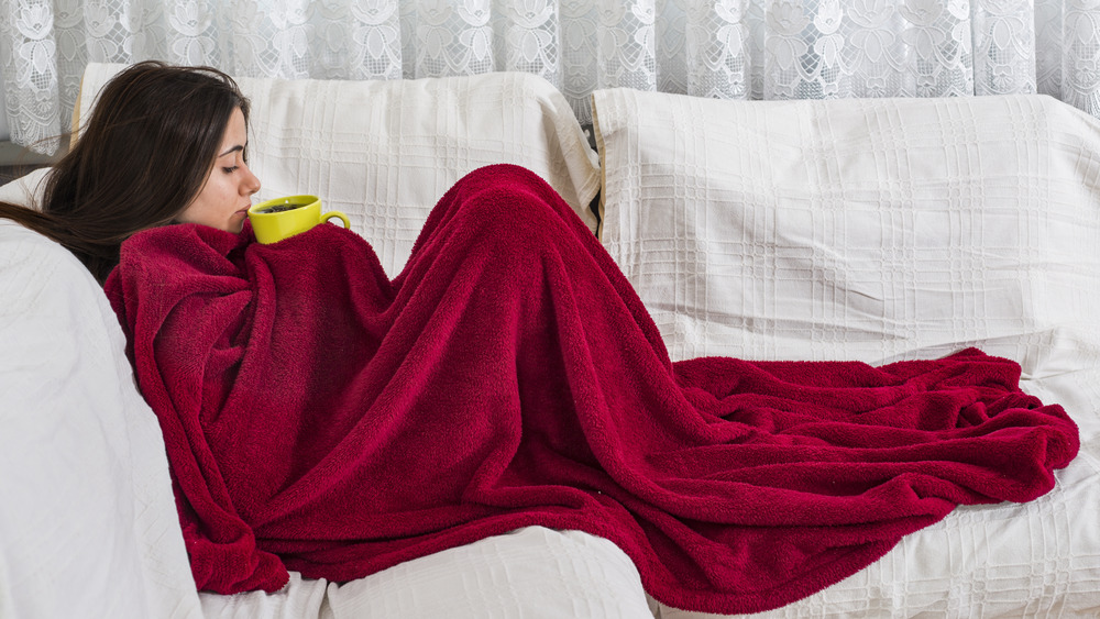 Person with long brown hair sits in profile on white couch wrapped in red blanket holding yellow mug