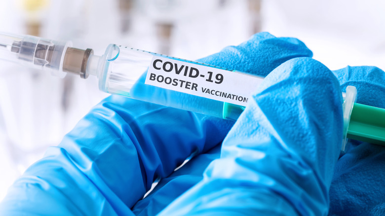 Gloved hand holding a syringe labeled "COVID-19 BOOSTER VACCINATION"