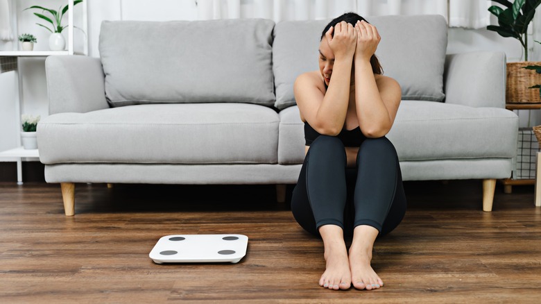 woman siting on floor next to scale looking stressed about weight