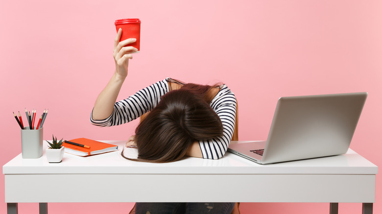 A woman with her head down on a desk as she holds up a red coffee travel cup