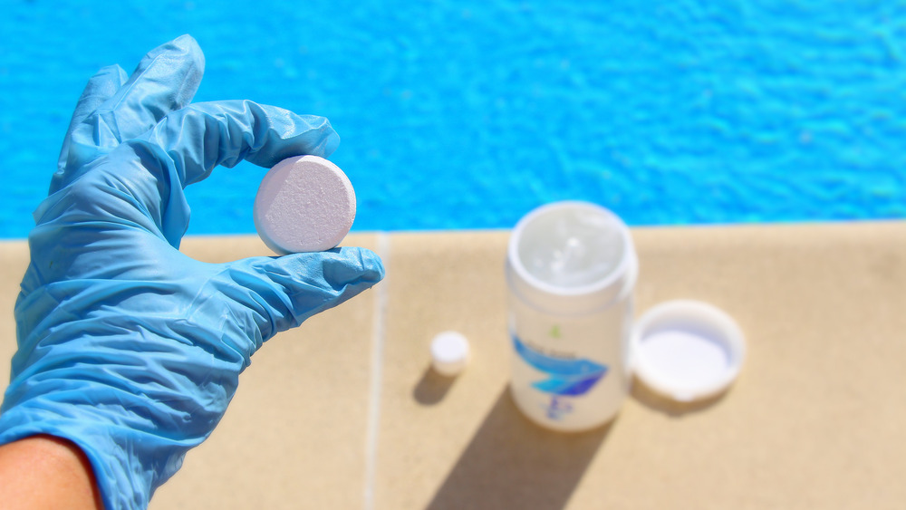 Gloved hand handling a chlorine tablet next to a pool.