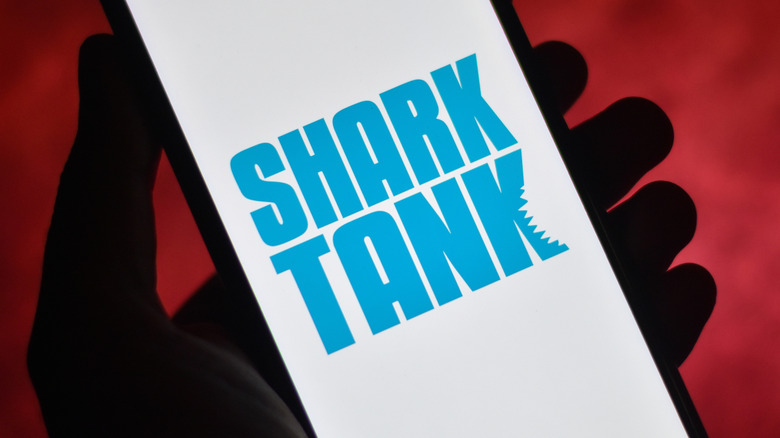 A hand holding a cell phone displaying the Shark Tank logo