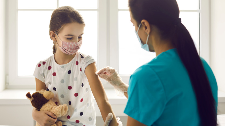 Young masked girl holding a stuffed animal receiving vaccine from masked doctor