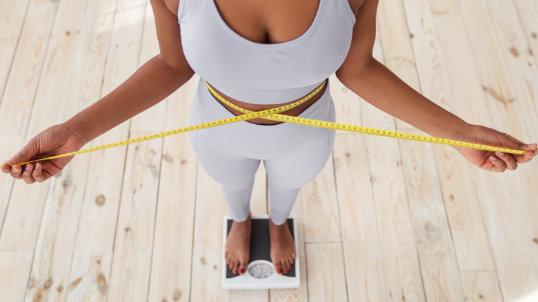 woman measuring her waist with tape while standing on a scale