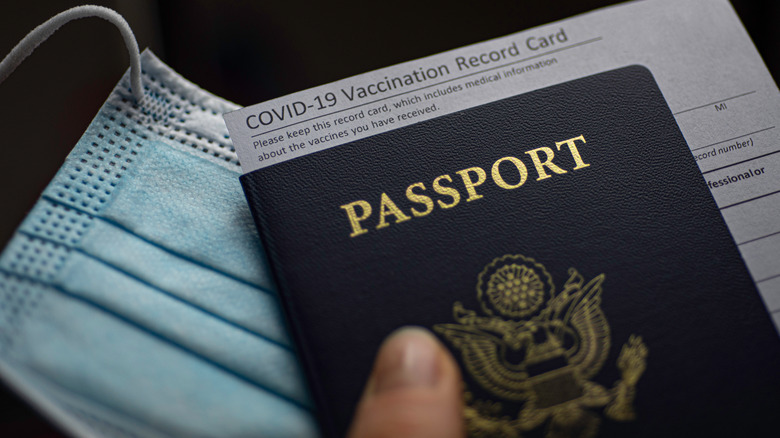 Close up of hand holding a blue passport and vaccination card next to a face mask