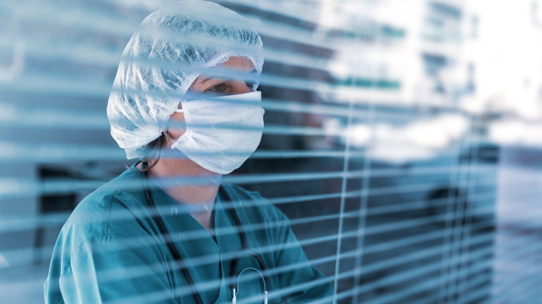 Health care worker in mask and hair net looking through window blinds