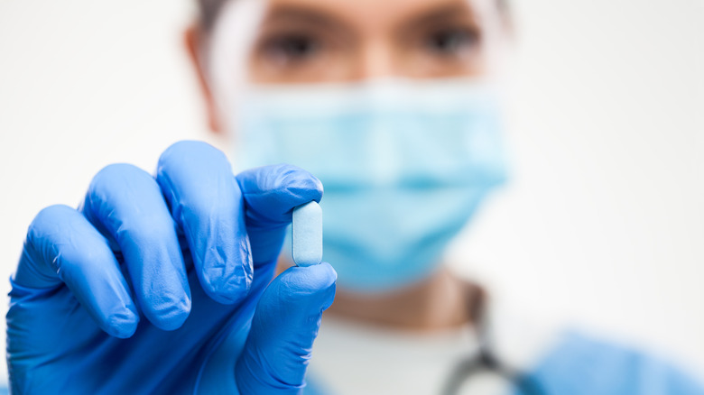 Doctor wearing protective face mask, gloves, holding blue pill, selective macro detail focus.