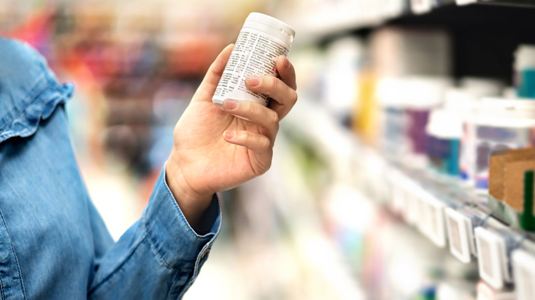 Person's hand holding a pill bottle with label showing while standing in the medicine aisle of a store