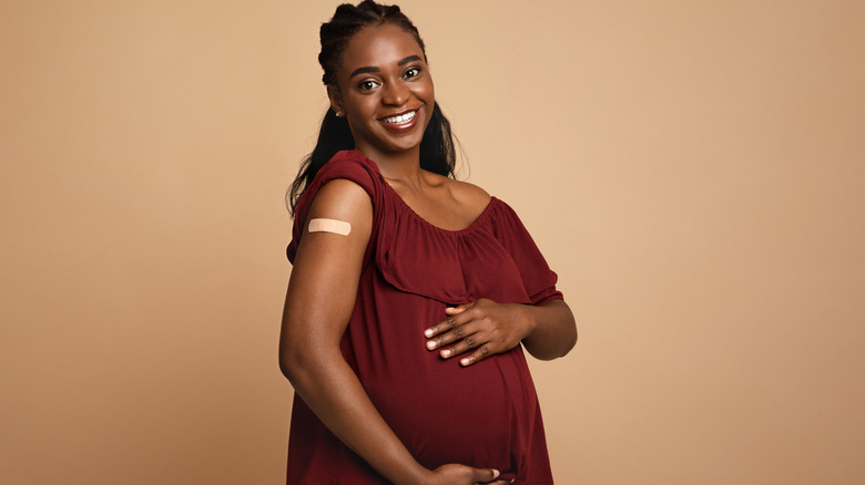 Woman smiling holding her pregnant belly with bandaid on her upper arm