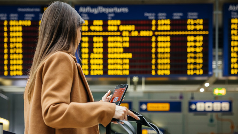 A woman checking her phone in front of an airport monitor