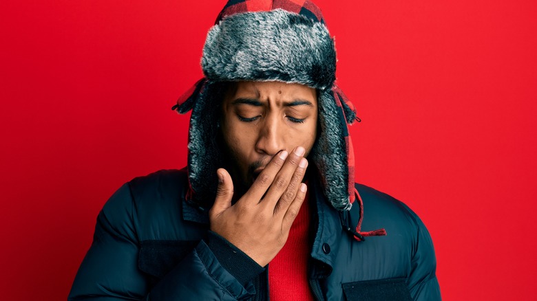 Man wearing a winter hat and coat yawning into his hand against a red background