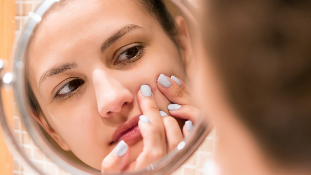 Young woman squeezes pimple on her face while looking in mirror