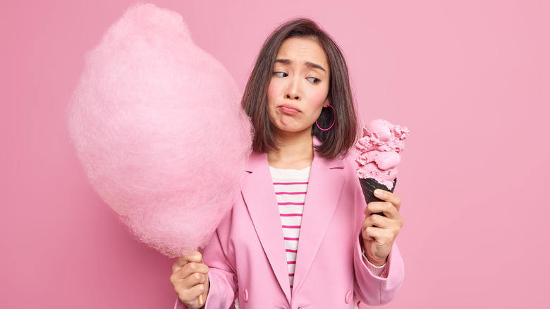 a young woman looks conflicted holding an ice cream cone and cotton candy in front of a pink backdrop