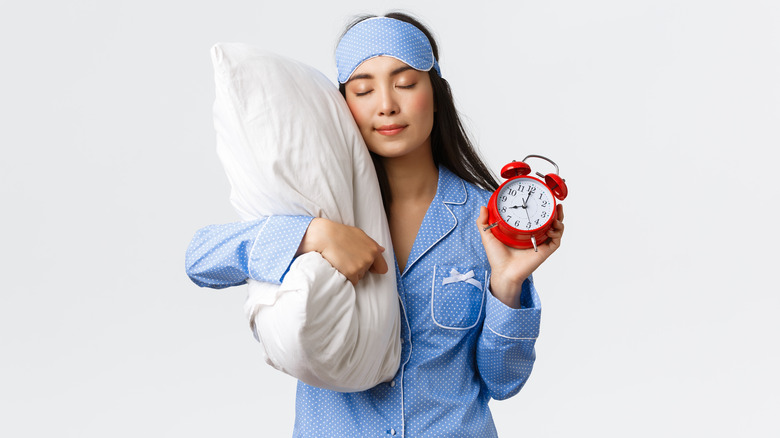 Woman in blue pajamas and sleep mask holding a pillow and alarm clock