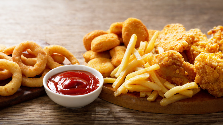 A small bowl of ketchup next to two wooden boards covered with onion rings, chicken nuggets, French fries, and fried chicken