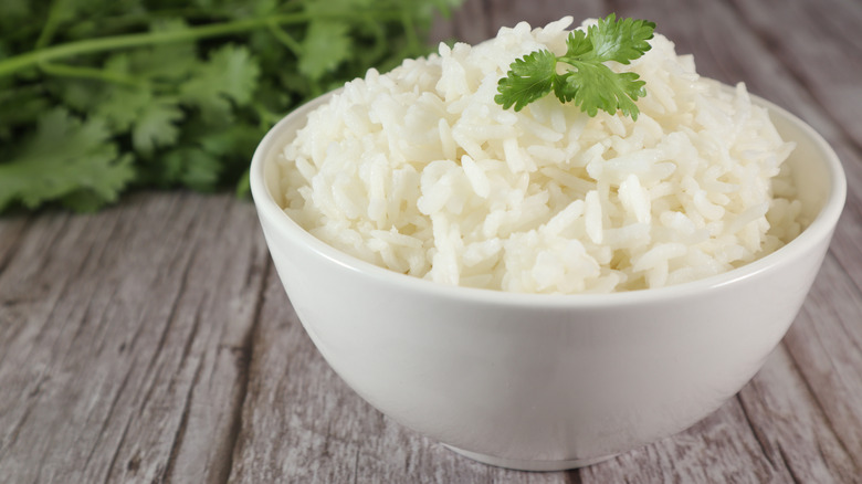 A bowl of white rice garnished with a sprig of parsley on a wooden surface with more parsley in the background