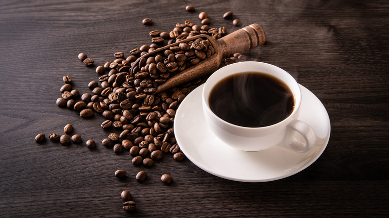 A cup of coffee on a saucer next to coffee beans and a wooden scoop on a wooden surface