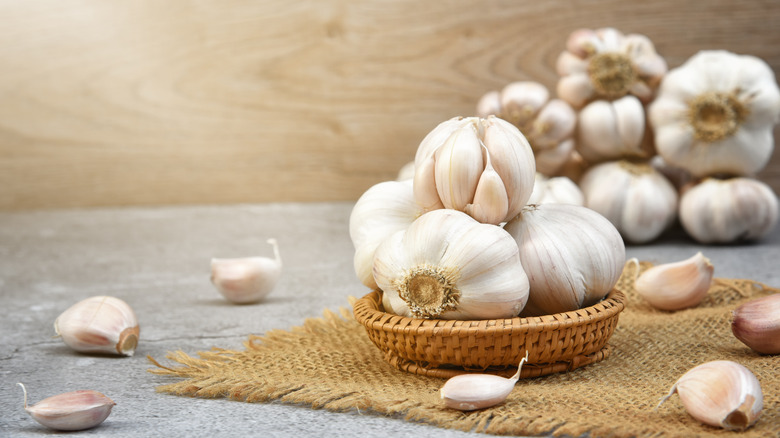 Garlic bulbs in a small wicker basket on burlap surrounded by garlic cloves with more garlic bulbs in the background