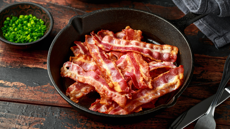 A skillet filled with bacon next to a small bowl of chives and a knife and fork