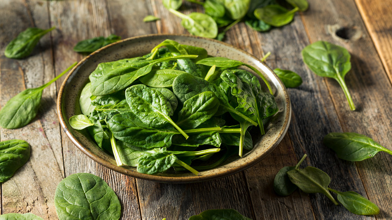 A bowl filled with spinach surrounded by spinach leaves on a wooden surface