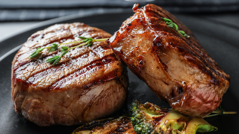 Two steaks with garnish next to broccoli on a dark gray plate