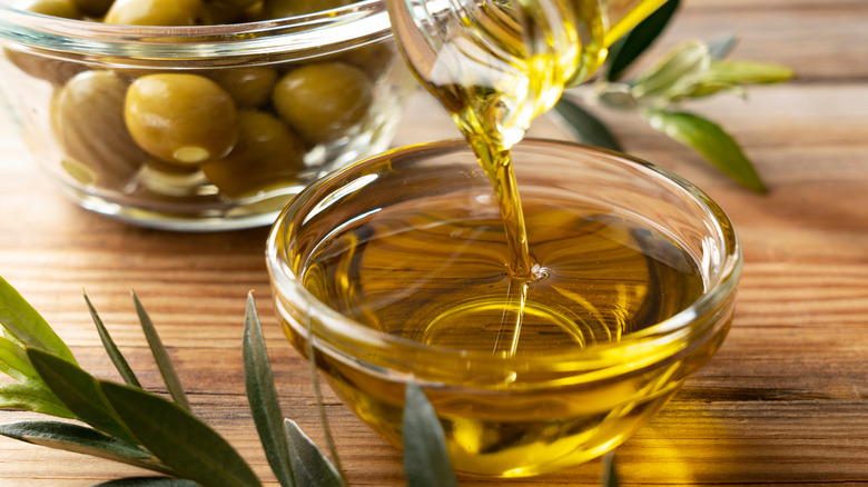 Close up of olive oil being poured from a glass bottle into a glass bowl with a bowl of green olives in the background