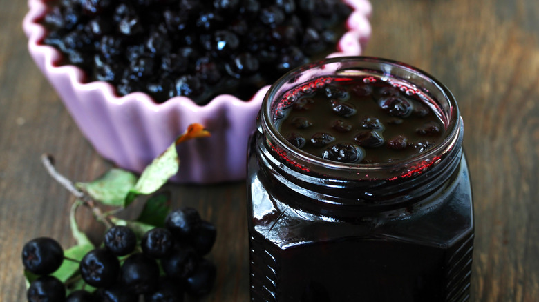 A jar of chokeberry jam next to a chokeberry branch and a lavender bowl filled with chokeberries