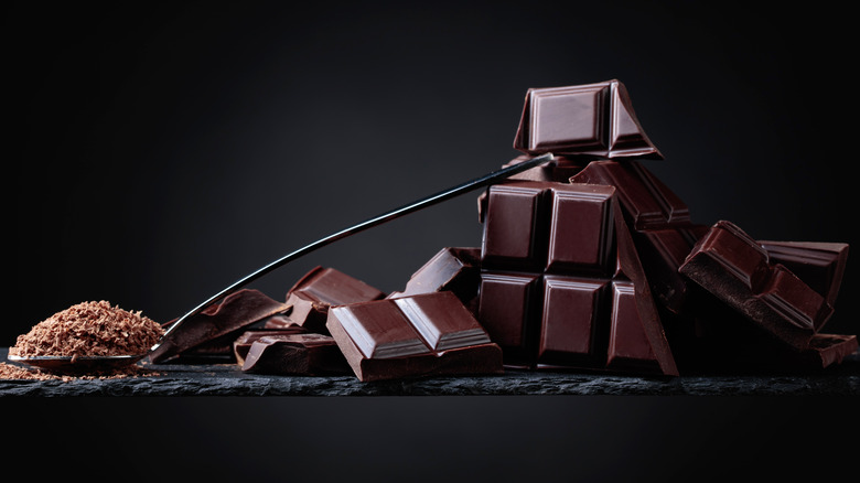 Pieces of squares of dark chocolate next to a spoon of bits of chocolate against a dark background