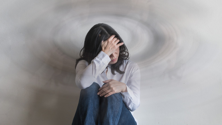 Woman sits on floor up against wall with her hand covering her facethat has concentric circles on it, indicating that she feels dizzy