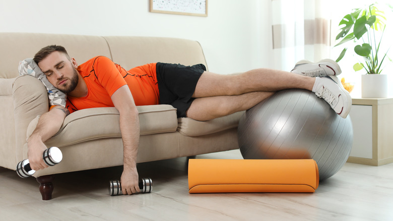 Man sleeping on the couch holding hand weights and resting his feet on a workout ball